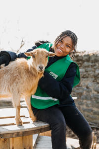A woman wearing a green vest sits on a table next to a goat