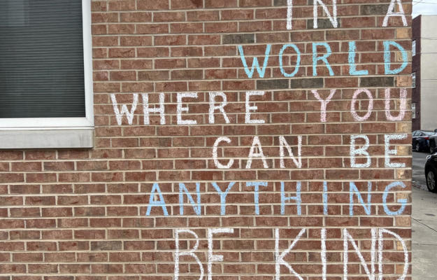 A brick wall with the words "in a world where you can be anything be kind" written on it.