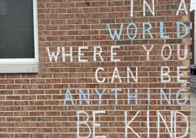 A brick wall with the words "in a world where you can be anything be kind" written on it.