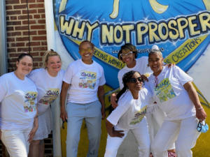 A group of women stand in front of a mural that reads Why Not Prosper
