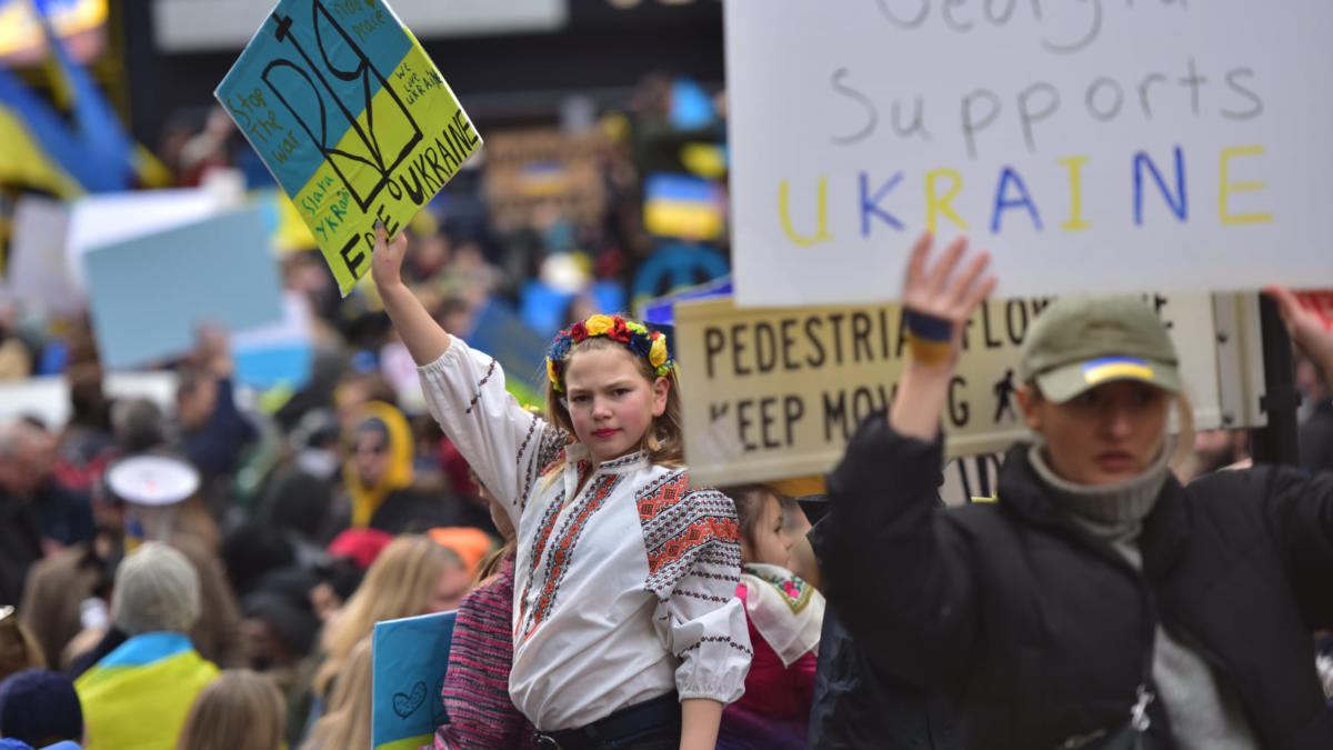 A woman in traditional Ukrainian garb holding a pro-Ukrainian sign defiantly while hundreds of protesters hold similar signs around her.