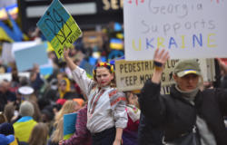 A woman in traditional Ukrainian garb holding a pro-Ukrainian sign defiantly while hundreds of protesters hold similar signs around her.