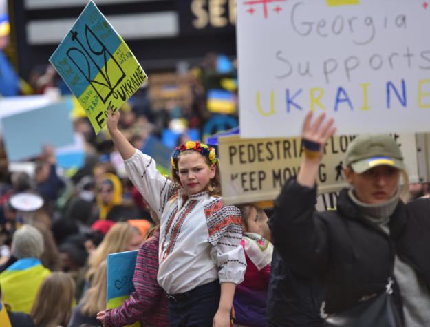 A woman in traditional Ukrainian garb holding a pro-Ukrainian sign defiantly while hundreds of protesters hold similar signs around her.