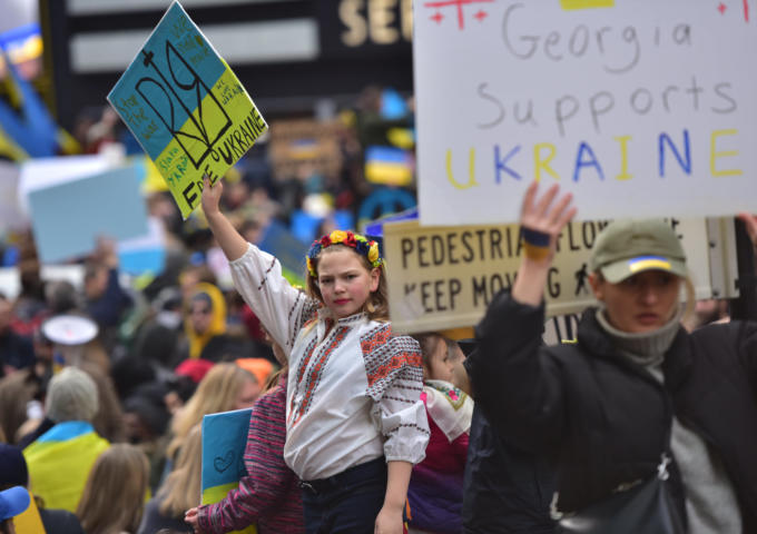 A woman in traditional Ukrainian garb holding a pro-Ukrainian sign defiantly while hundreds of protesters hold similar signs around her.