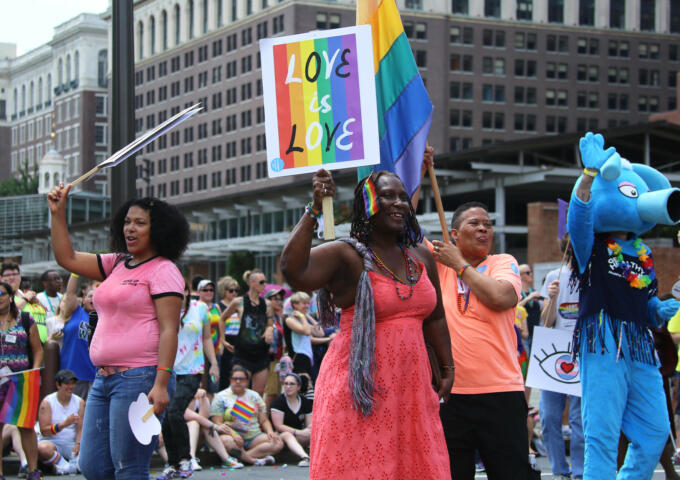 A diverse group of LGBTQ community members, including cisgender and transgender people, Black people, white people, and others holding rainbow signs and messages in front of Independent Hall in Philadelphia.