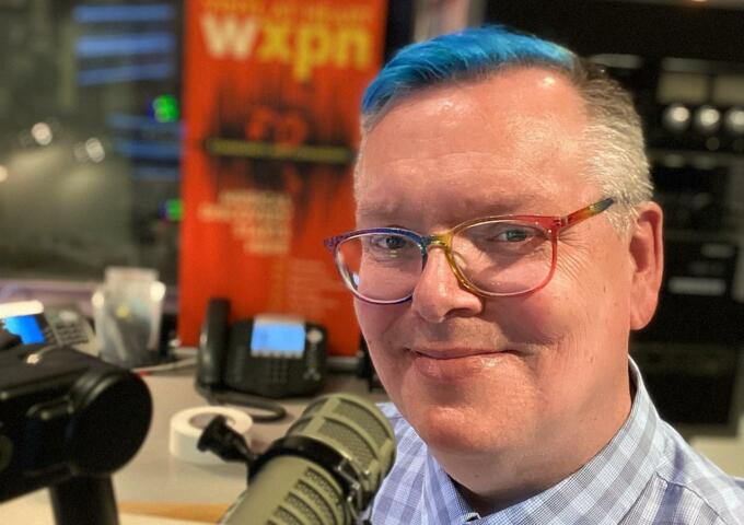 DJ Robert Drake in the WXPN studio smiling with a professional microphone near his mouth. He is wearing glasses.