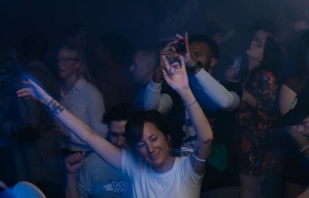 A smiling woman dancing with her hands in the air with a crowd behind her at a Shakedown party.