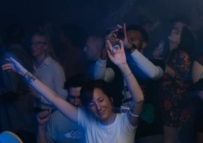 A smiling woman dancing with her hands in the air with a crowd behind her at a Shakedown party.