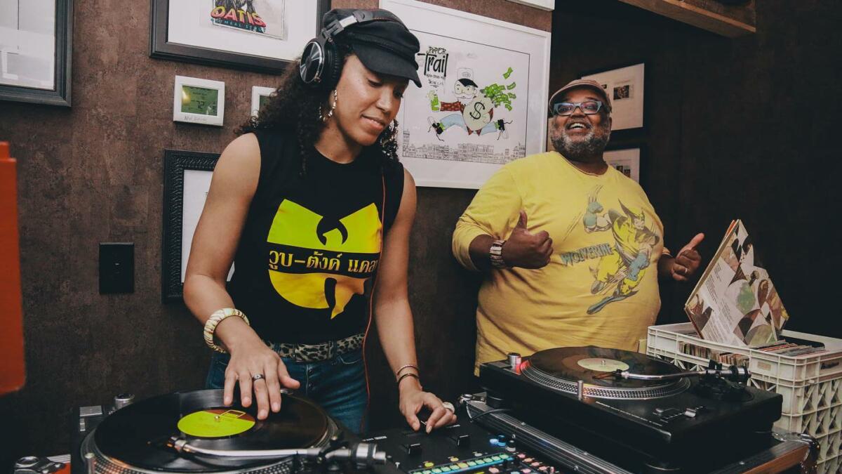 A woman with a Wu-Tang Clan t-shirt spins a record at a turntable while DuiJi Mschinda smiles at the crowd as he stands next to her.