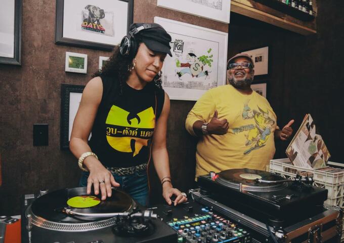 A woman with a Wu-Tang Clan t-shirt spins a record at a turntable while DuiJi Mschinda smiles at the crowd as he stands next to her.